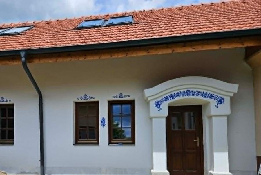 Bílá dvorní přístavba s červenou střechou, okny a portálem zdobenými modrými ornamenty