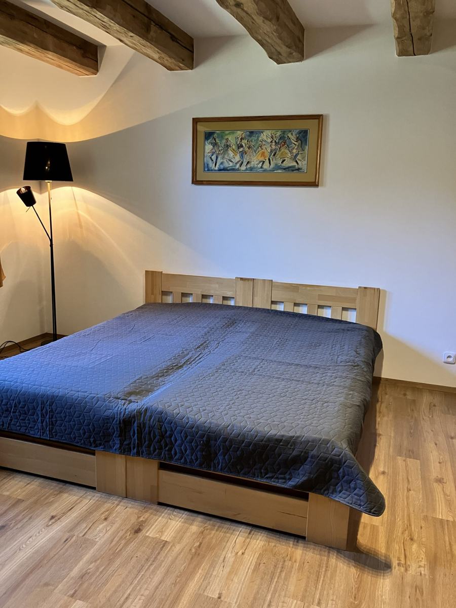 Postel s modrým přehozem, nad ní obraz s lidovým motivem a stojací lampa